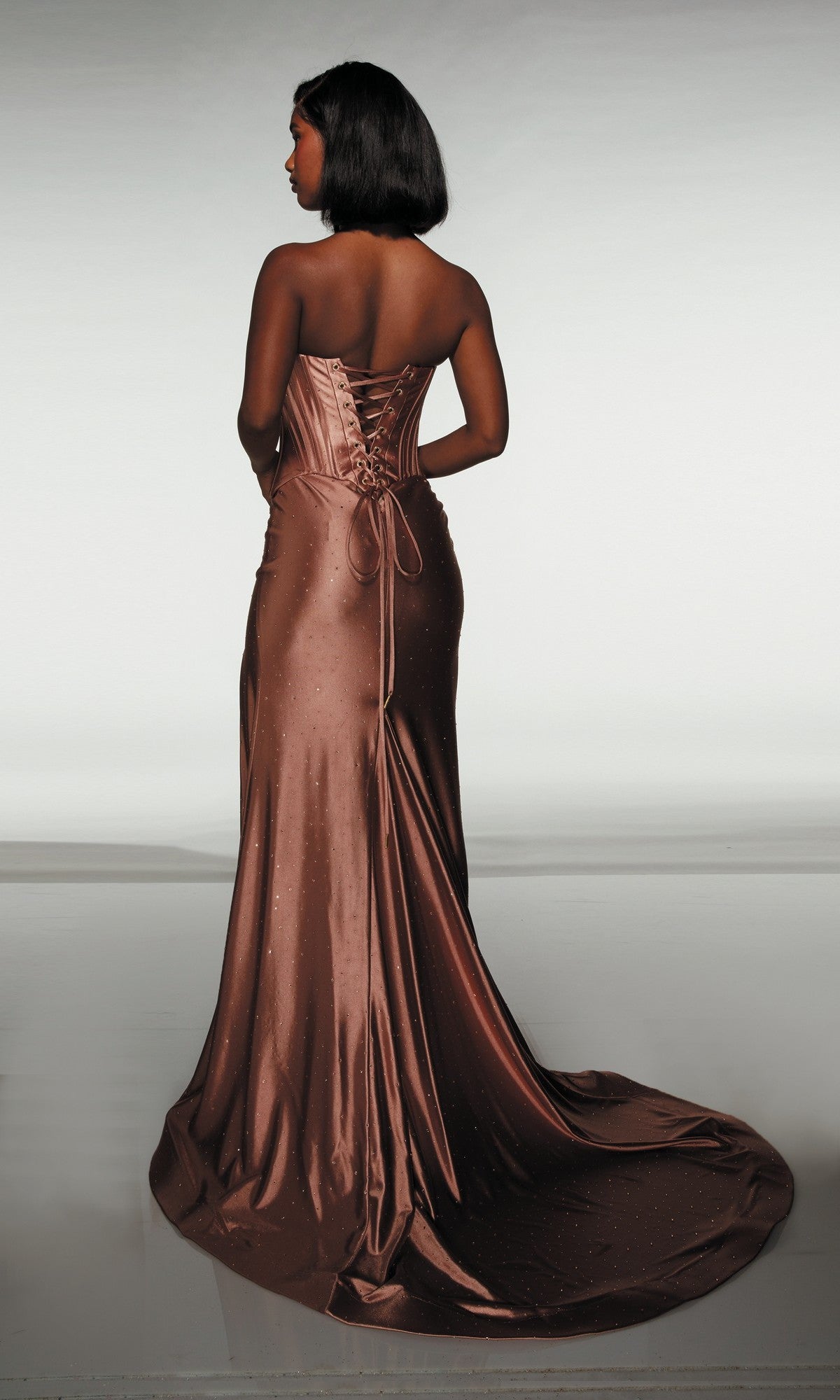 A woman faces away from the camera in the Alyce 62094 long prom dress, a brown satin strapless gown with hot stone embellishments, lace-up back, and long train, standing against a plain background.